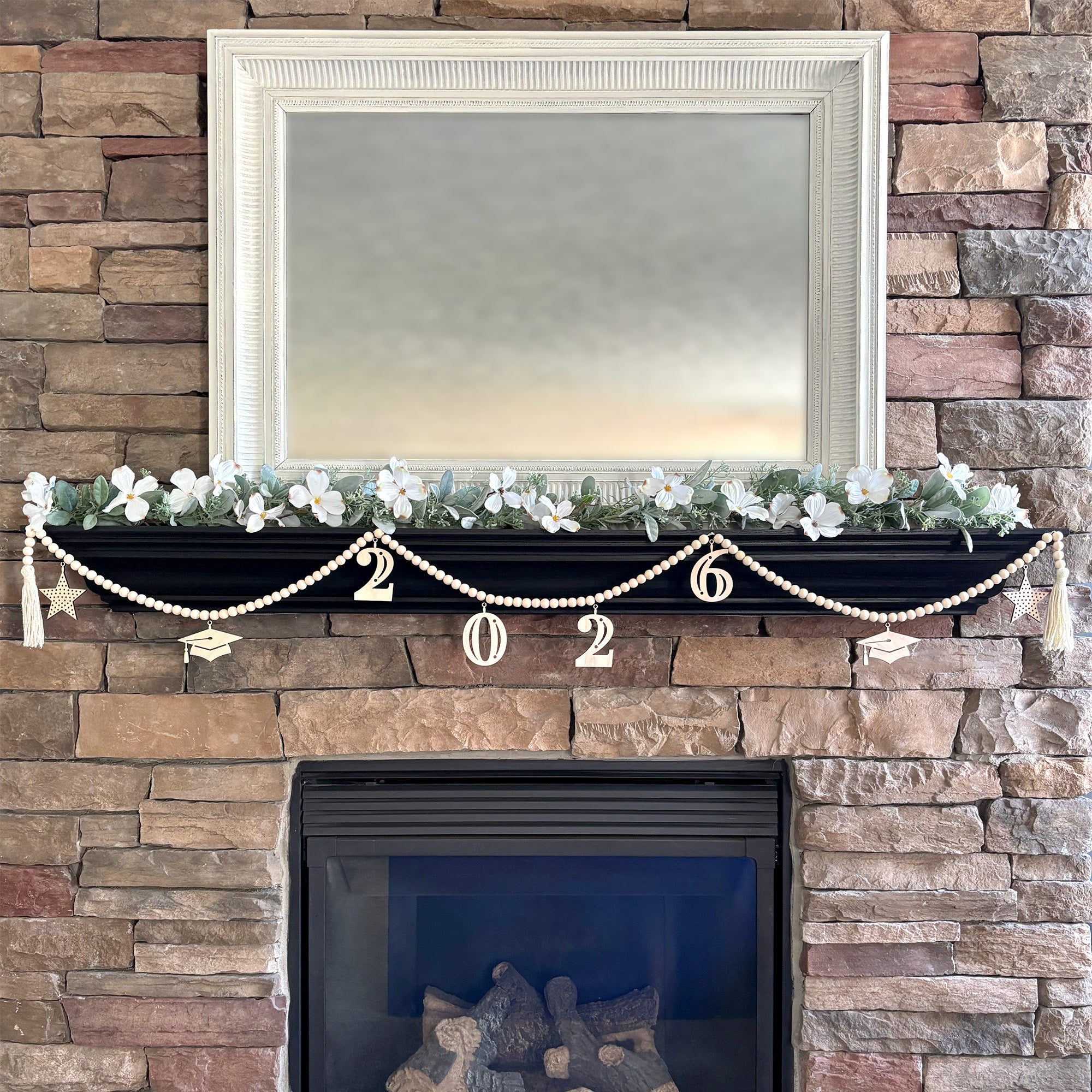 2026 Graduation decoration on fireplace mantel. Garland is made from wood beads with laser cut wood charms that read 2026. Additional charms are mortar board hats and stars.